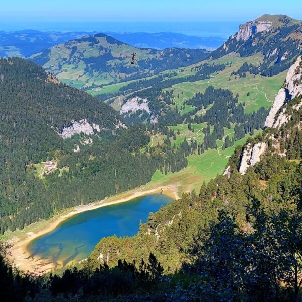 Ein Panoramablick auf ein üppig grünes Tal mit einem kleinen See in seiner Mitte, umgeben von bewaldeten Hügeln und felsigen Bergen unter einem klaren blauen Himmel.