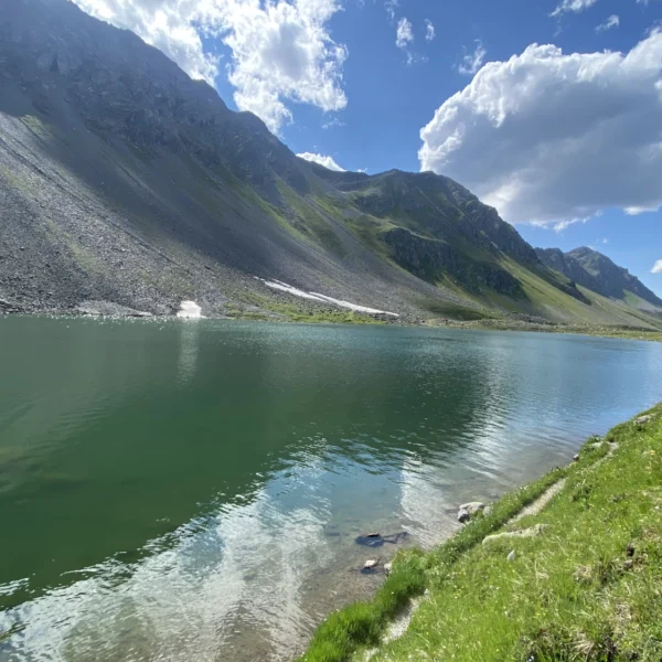Ein klarer Bergsee wie der Schottensee spiegelt blauen Himmel und Wolken wider, umgeben von steilen Felshängen und grünem Gras im hellen Sonnenlicht.
