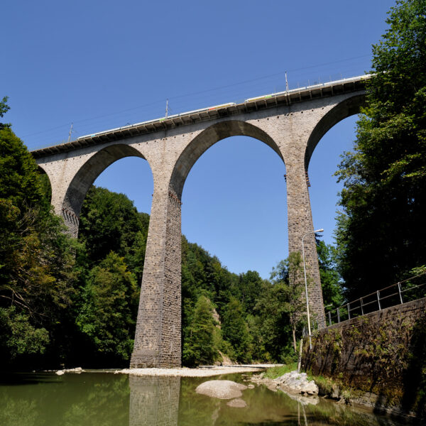 Hohes Steinviadukt über den Fluss, umgeben von grünen Bäumen.