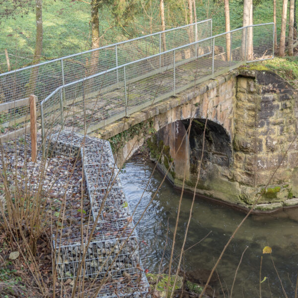 Steinbrücke mit Metallgeländer über einen kleinen Fluss. Uferbefestigung mit Steinkörben.