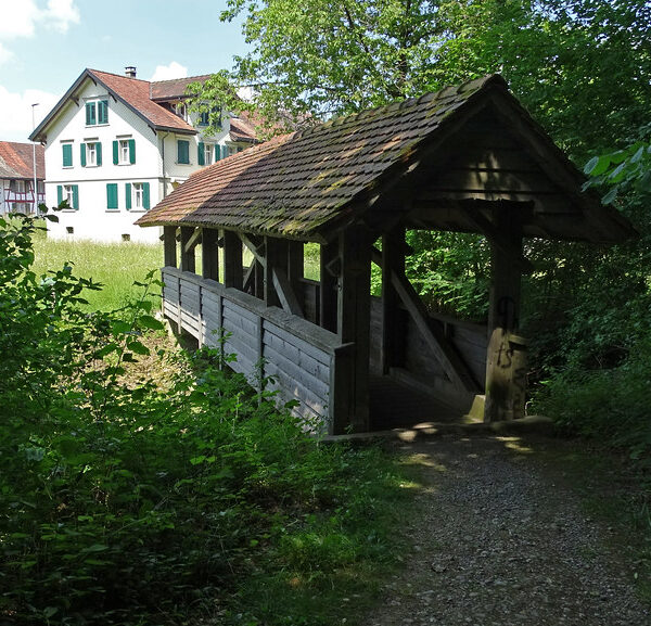 Überdachte Holzbrücke über den Tanneggerbach, umgeben von Grün.