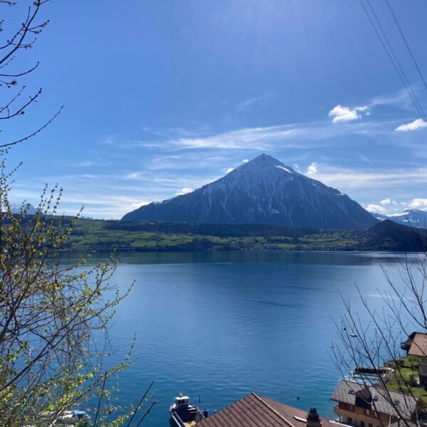 Ein schneebedeckter Berg erhebt sich über einem blauen See, über dem ein klarer Himmel liegt. Im Vordergrund sind Dächer, Boote und Baumzweige zu sehen, die auf ein friedliches Dorf am See hinweisen.