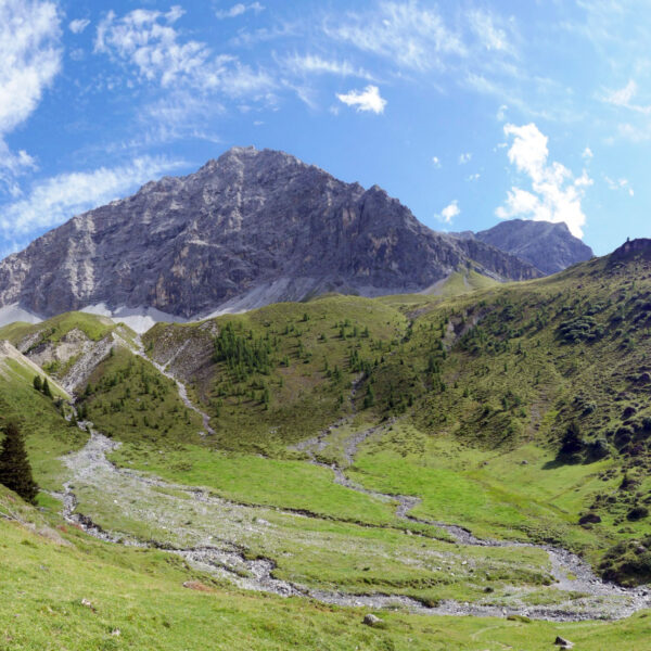 Tiejerbach: Berglandschaft mit grünen Wiesen und felsigem Gipfel unter blauem Himmel.