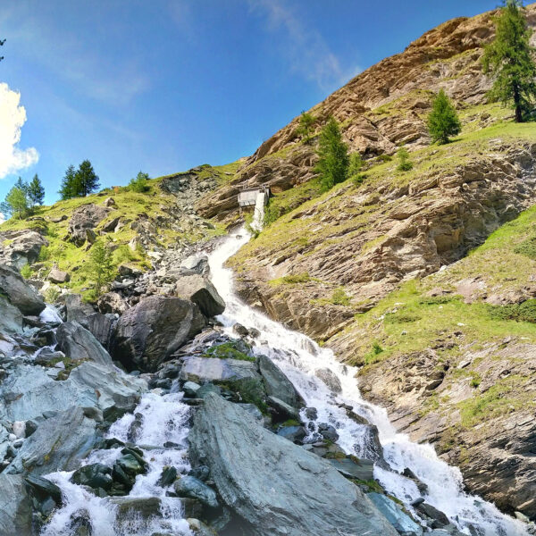 Triftbach Wasserfall in Zermatt mit Felsen und grüner Vegetation unter blauem Himmel.