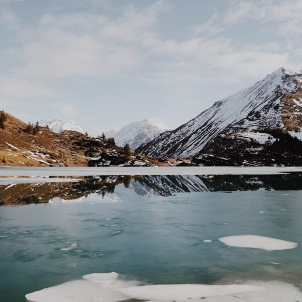 Teilweise zugefrorener See mit schwimmendem Eis, umgeben von schneebedeckten Bergen und braunen Hügeln, die sich im ruhigen Wasser unter einem teilweise bewölkten Himmel spiegeln.