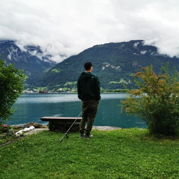 Eine Person steht auf einer Wiese mit Blick auf einen ruhigen See. In der Nähe liegt eine Angelrute auf dem Boden. Dahinter zeichnen sich neblige Berge und ein bewölkter Himmel ab, die eine friedliche und malerische Landschaft schaffen.
