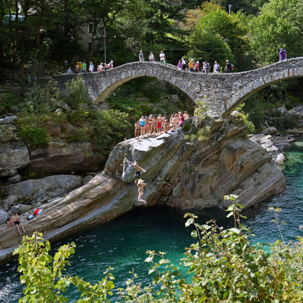 Steinbrücke in Verzasca, Schweiz, mit Menschen beim Klippenspringen in türkisfarbenes Wasser.