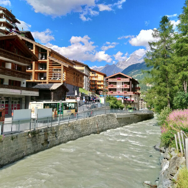 Vispa Fluss in Zermatt, Schweiz, mit traditionellen Holzhäusern und Bergen im Hintergrund. Gemeinde Zermatt Schild sichtbar.
