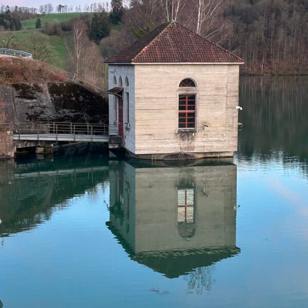 Ein kleines, quadratisches Gebäude mit gewölbten Fenstern liegt teilweise im ruhigen Wasser und spiegelt sein Bild wider. Im Hintergrund stehen Bäume ohne Blätter und ein grasbewachsener Hügel unter einem blau-rosa Himmel.