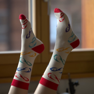 Die Füße einer Person stehen vor einem Fenster und tragen 1 Paar Angeln-Fischen.ch Socken: cremefarben mit roten Zehen, Fersen und Bündchen sowie bunten Fischmustern - perfekt für Angelfans.