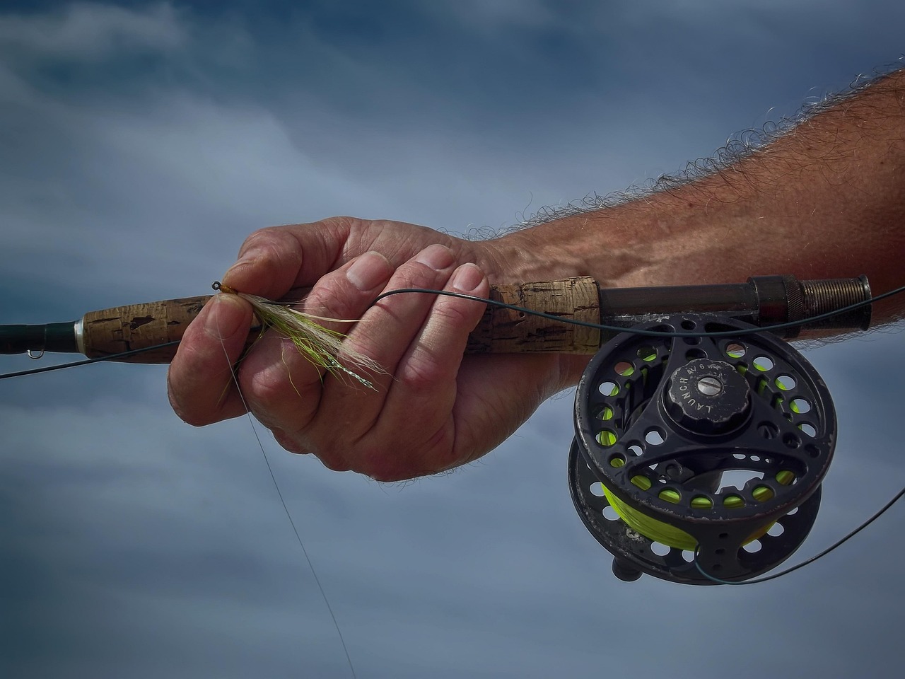 Hand hält Fliegenfischerrute mit Rolle und Fliege. Ausrüstung zum Fliegenfischen in der Schweiz.