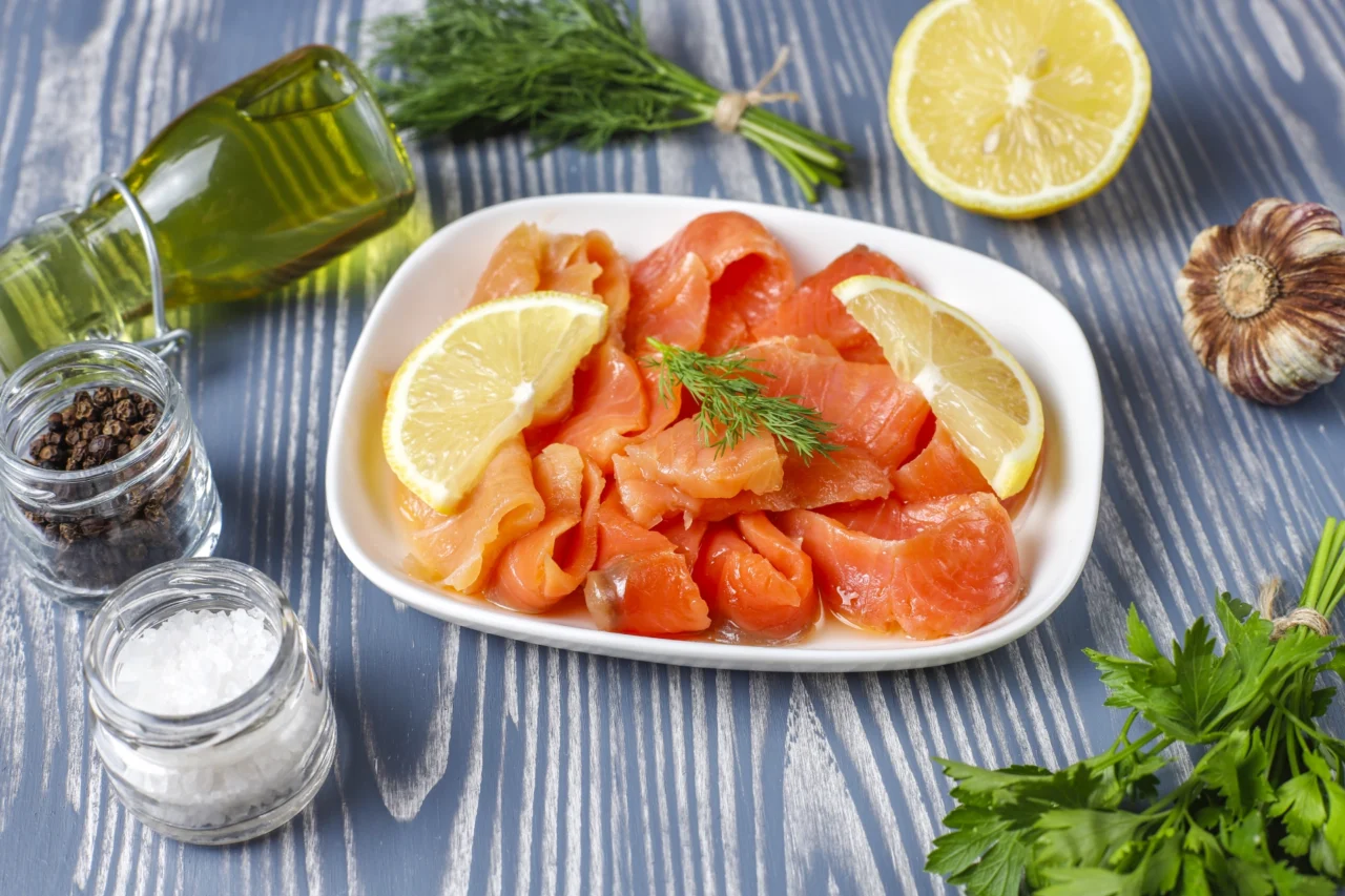 Ein Teller mit Räucherlachs, garniert mit Zitronenscheiben und Dill, umgeben von Petersilie, Knoblauch, einer halbierten Zitrone, einer Flasche Olivenöl und kleinen Gläsern mit Salz und Pfeffer auf einer Holzplatte - ein Geschmack, der an Kanada erinnert.