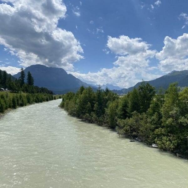 Ein breiter, blasser Fluss fließt zwischen dichten grünen Bäumen mit Bergen und einem bewölkten blauen Himmel im Hintergrund.
