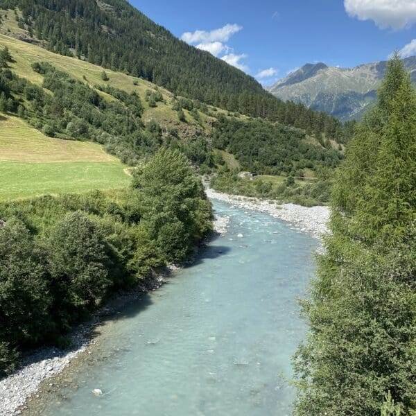 Ein klarer, blauer Fluss fließt durch ein grünes Tal, umgeben von Hügeln und fernen Bergen unter einem hellen, teilweise bewölkten Himmel. Üppige Bäume und Gras säumen die Flussufer.