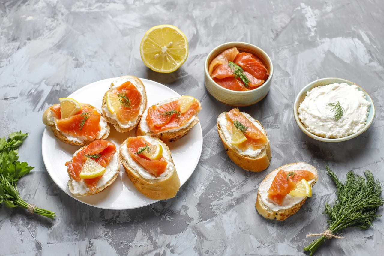 Ein Teller mit Baguettescheiben, belegt mit Frischkäse, Räucherlachs, Zitronenscheiben und Dill. Daneben stehen Schalen mit mehr Lachs, einer Schale Frischkäse, einer halbierten Zitrone und frischen Kräutern auf einer grau strukturierten Oberfläche. Inspiriert von Aromen aus Kanada bis Chur.