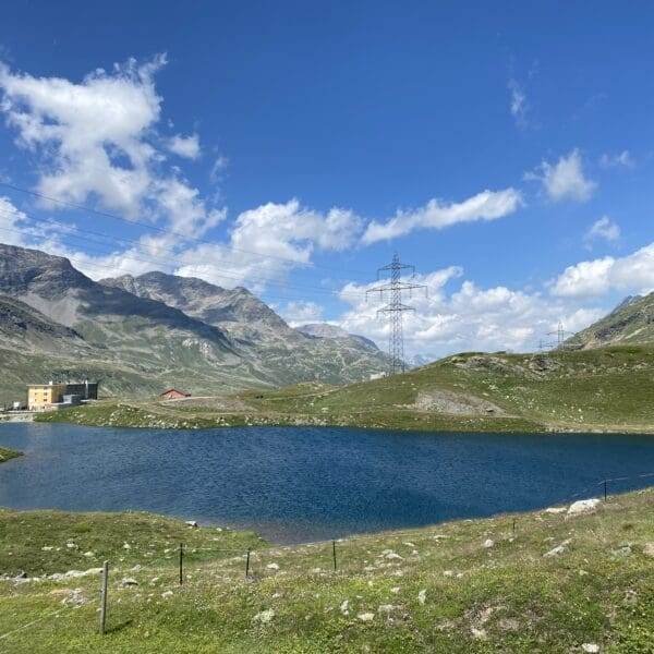 Ein kleiner See, der Lagh da la Cruseta, umgeben von grünen Hügeln und Bergen unter einem blauen, teilweise bewölkten Himmel in der Nähe des Berninapasses. Ein Gebäude und ein roter Schuppen stehen am Wasser, Stromleitungen und Masten durchziehen die Landschaft.