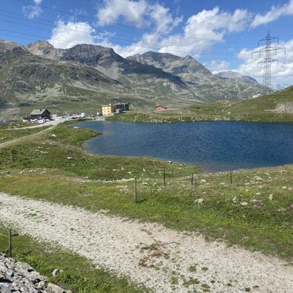 Eine ruhige Berglandschaft in der Nähe des Lagh da la Cruseta am Berninapass mit einem klaren blauen See, Grasfeldern, einem Feldweg, Gebäuden in der Ferne und Bergen unter blauem Himmel. Rechts sind eine Stromleitung und ein Strommast zu sehen.