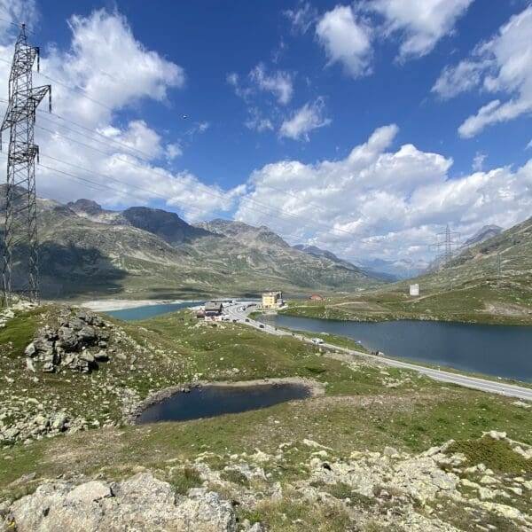 Eine malerische Berglandschaft entlang des Berninapasses mit einer kurvenreichen Straße, einem kleinen See (Lagh da la Cruseta), felsigen Hügeln, Grasfeldern, einem gelben Gebäude und Stromleitungen unter einem teilweise bewölkten blauen Himmel.