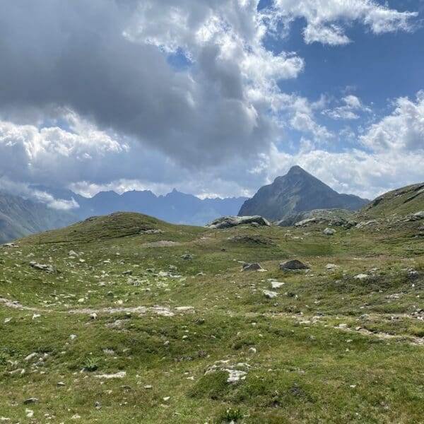 Grasbewachsene Hügel und felsiges Gelände unter einem teilweise bewölkten Himmel in der Nähe des Berninapasses, mit einem dramatischen Berggipfel in der Ferne und Sonnenlicht, das durch die Wolken bricht.
