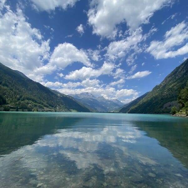 Klarer See, umgeben von grünen Bergen, in denen sich der teilweise bewölkte blaue Himmel spiegelt. Die fernen Alpen und Wolken bilden eine ruhige, malerische Landschaft am Lago di Poschiavo. In Ufernähe sind Felsen unter Wasser sichtbar.