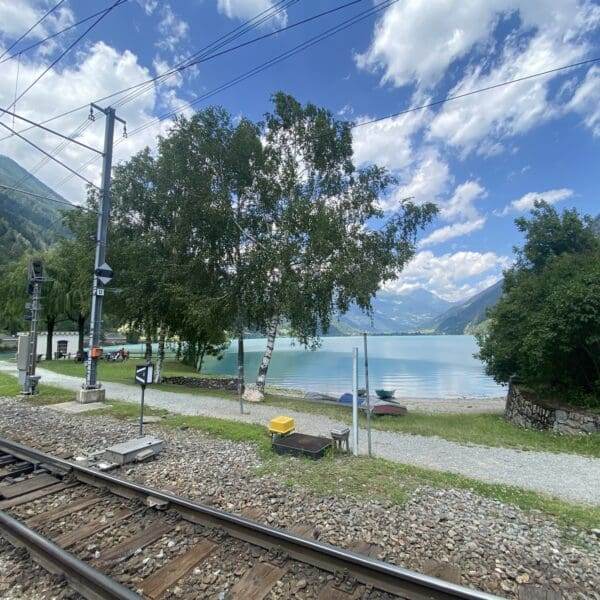 Ein malerischer Blick auf den Poschiavo-See mit türkisfarbenem Wasser, Bergen im Hintergrund, einem Baum in der Mitte und Eisenbahnschienen im Vordergrund unter einem teilweise bewölkten Himmel.