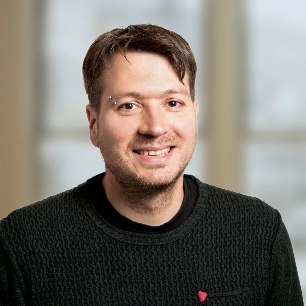 Ein lächelnder Mann mit kurzen braunen Haaren und einem Piercing im Gesicht trägt einen dunklen strukturierten Pullover und sitzt vor einem unscharfen Innenhintergrund mit großen Fenstern.