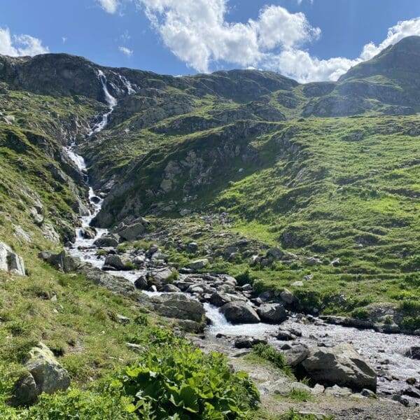 Berglandschaft mit einem kleinen Wasserfall und einem Bach, bekannt als Ova da Radönt, der unter einem teilweise bewölkten blauen Himmel grasbewachsene, felsige Hänge hinabfließt. Sonnenlicht hebt die grünen Hügel und verstreuten Felsen entlang des Wasserlaufs hervor.