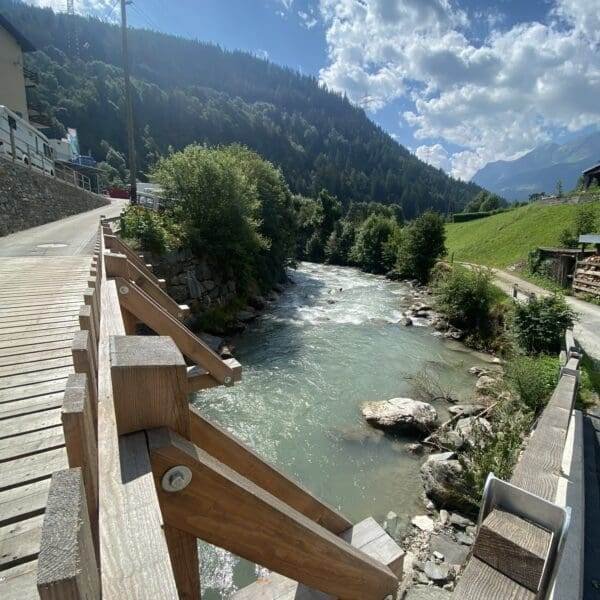 Eine Holzbrücke überquert einen klaren, fließenden Fluss, umgeben von grünen Bäumen, felsigen Ufern und grasbewachsenen Hügeln, mit Bergen und einem teilweise bewölkten Himmel im Hintergrund.
